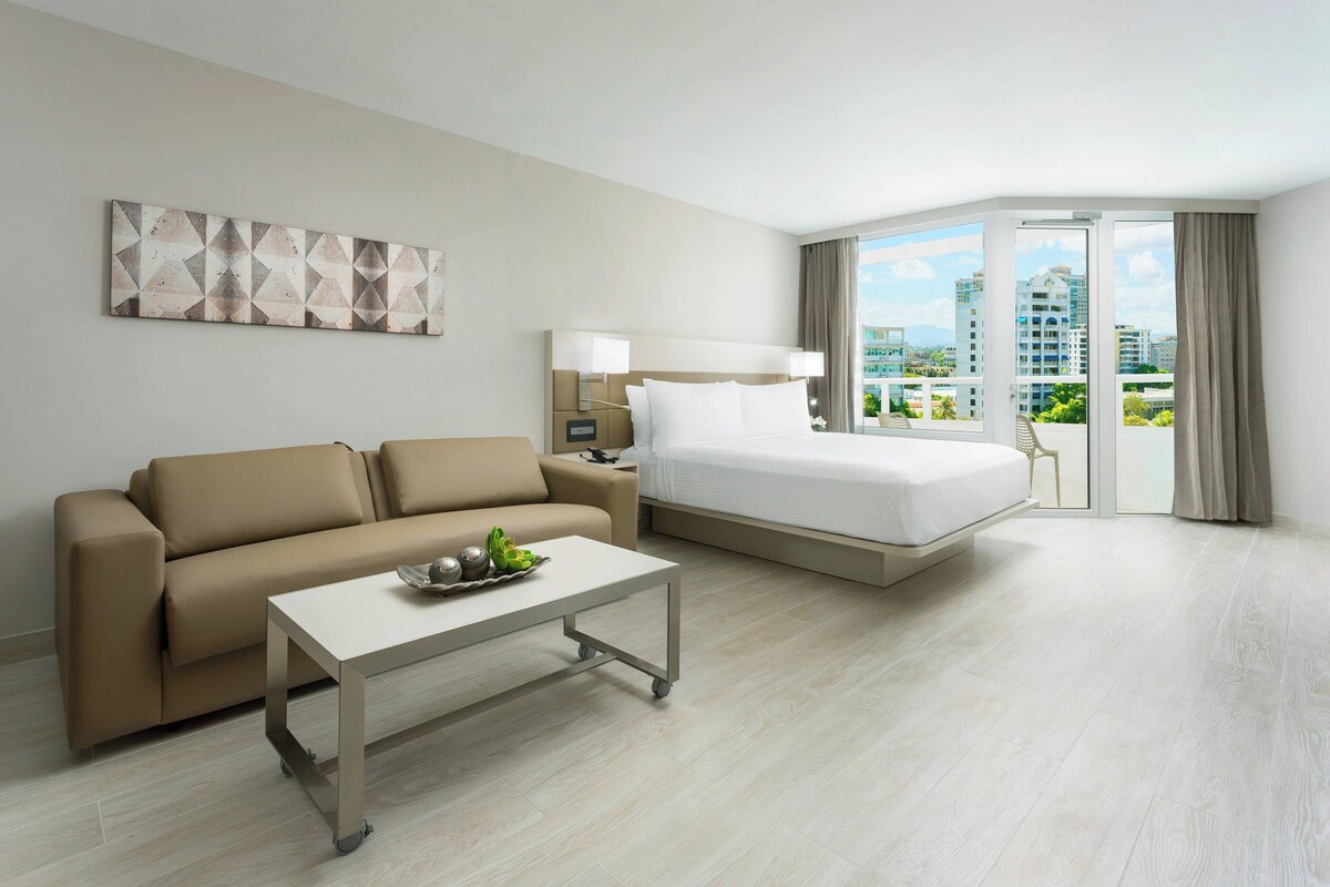 A hotel room with a bed, a sofa, a small table, and a view of the city through large windows.