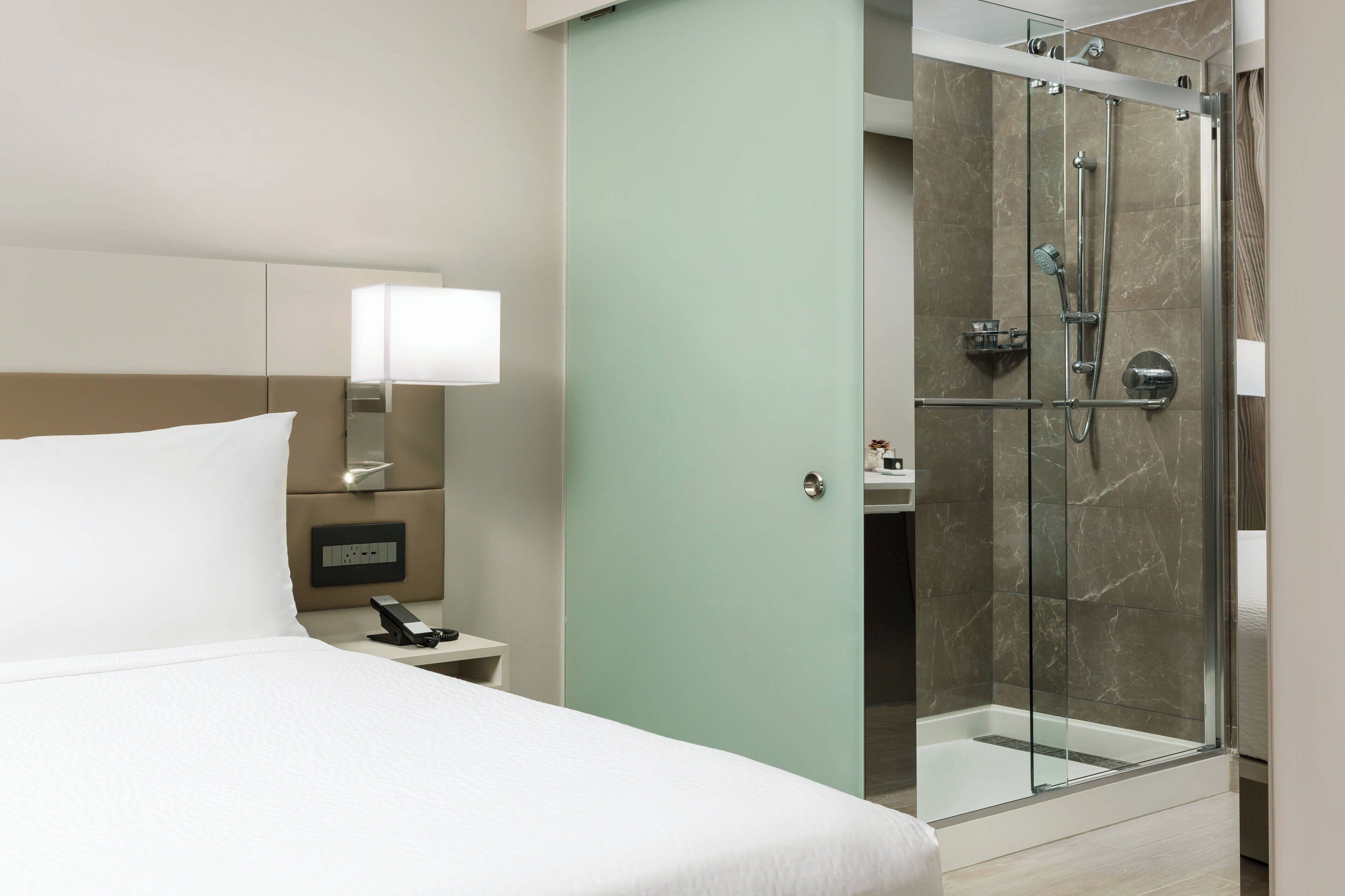 Habitación de hotel con cama, mesita de noche, lámpara y zona de ducha con mampara de vidrio.