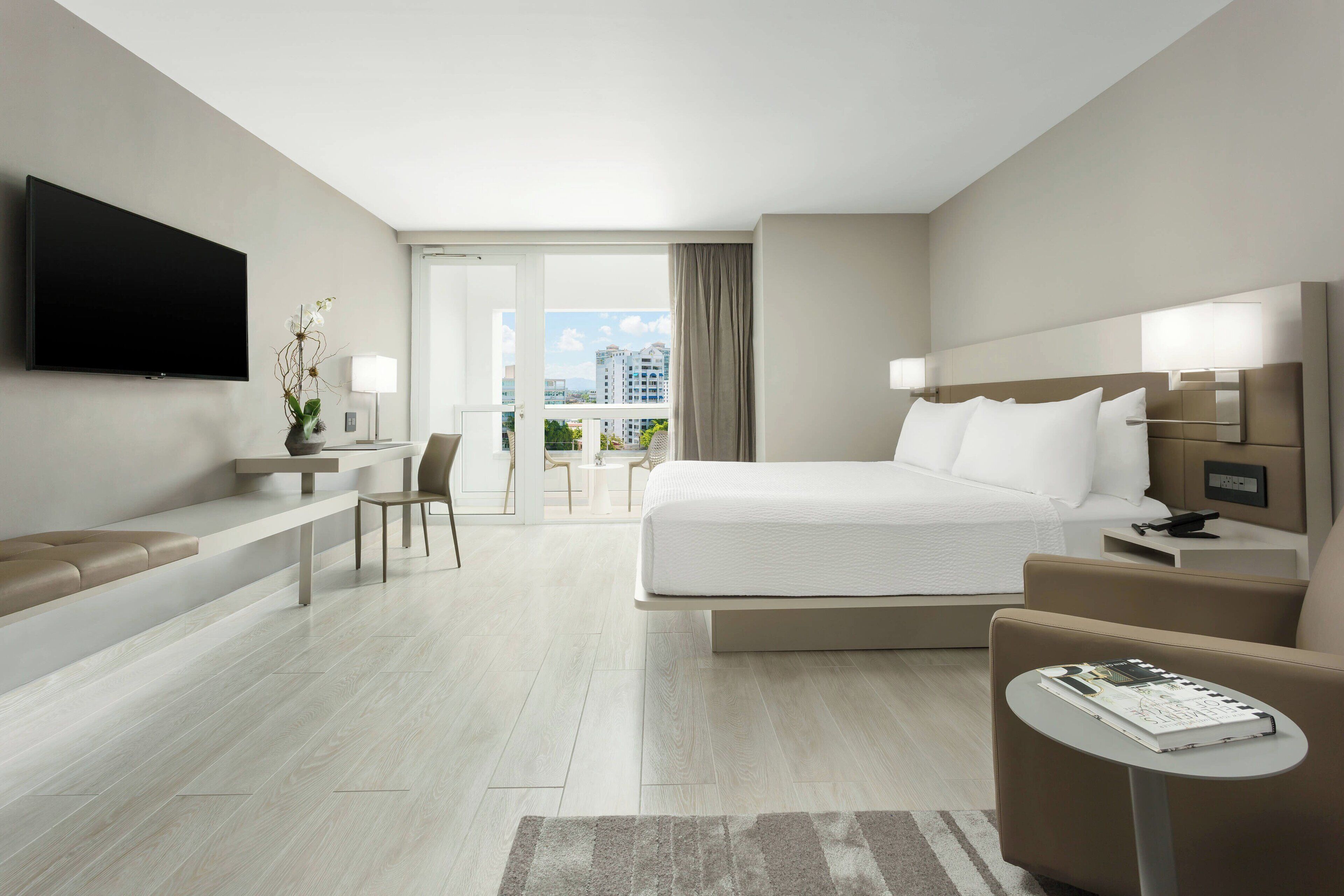 A modern hotel room with a large bed, a seating area, a TV, and a balcony with a view of the city.