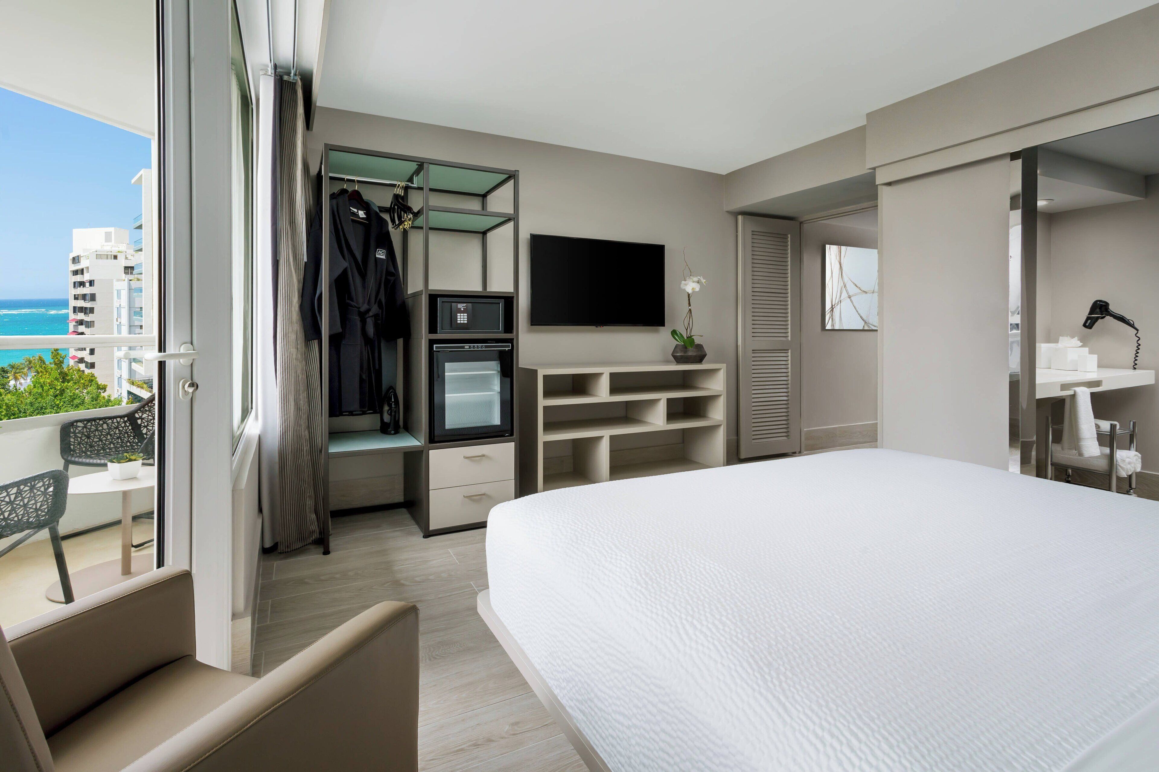 A modern hotel room with a large bed, a TV, a minibar, a seating area with a view of the ocean, and a balcony with a chair.