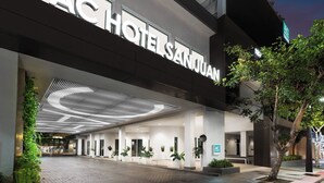 Fachada de hotel con diseño moderno, destacando un gran letrero iluminado, un área de acceso cubierta y plantas en macetas.