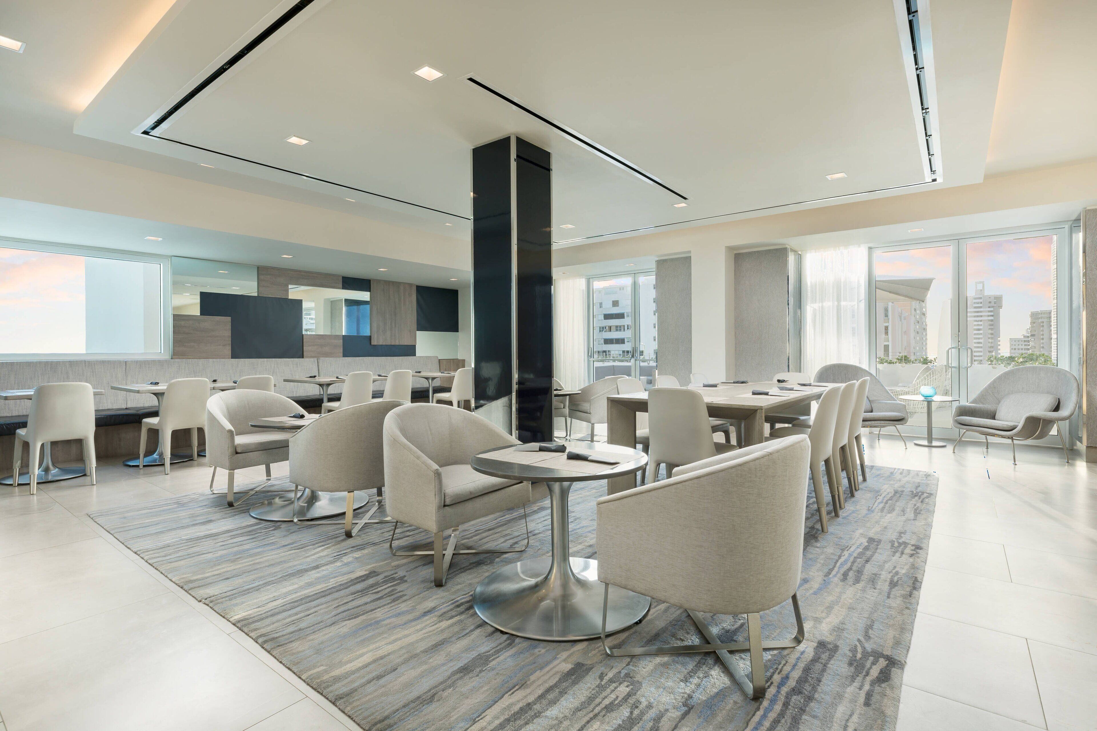 Un espace de restauration moderne avec des tables et des chaises rondes, un pilier central et de grandes fenêtres offrant une vue sur la ville.