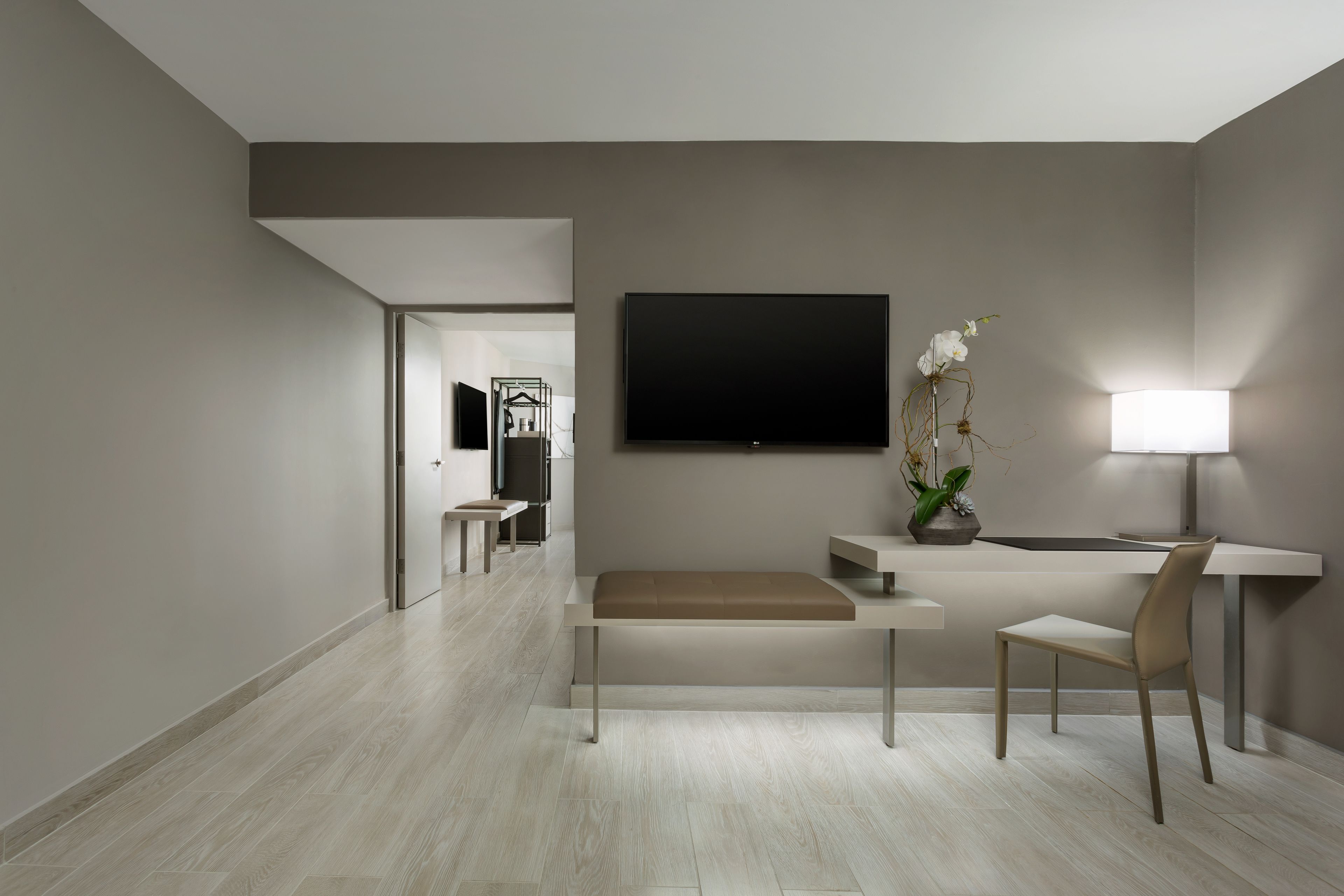 A modern hotel room with a flat-screen TV, a desk with a chair, a bench, and a bedside table.