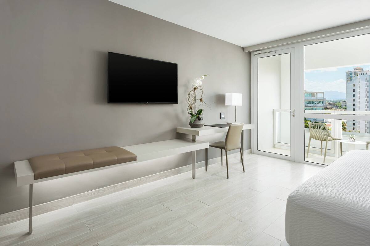 Ein modernes Hotelzimmer mit großem Flachbildfernseher, einer Bank, einem Schreibtisch mit Stuhl, einer Topfpflanze und einem Balkon mit Stadtblick.