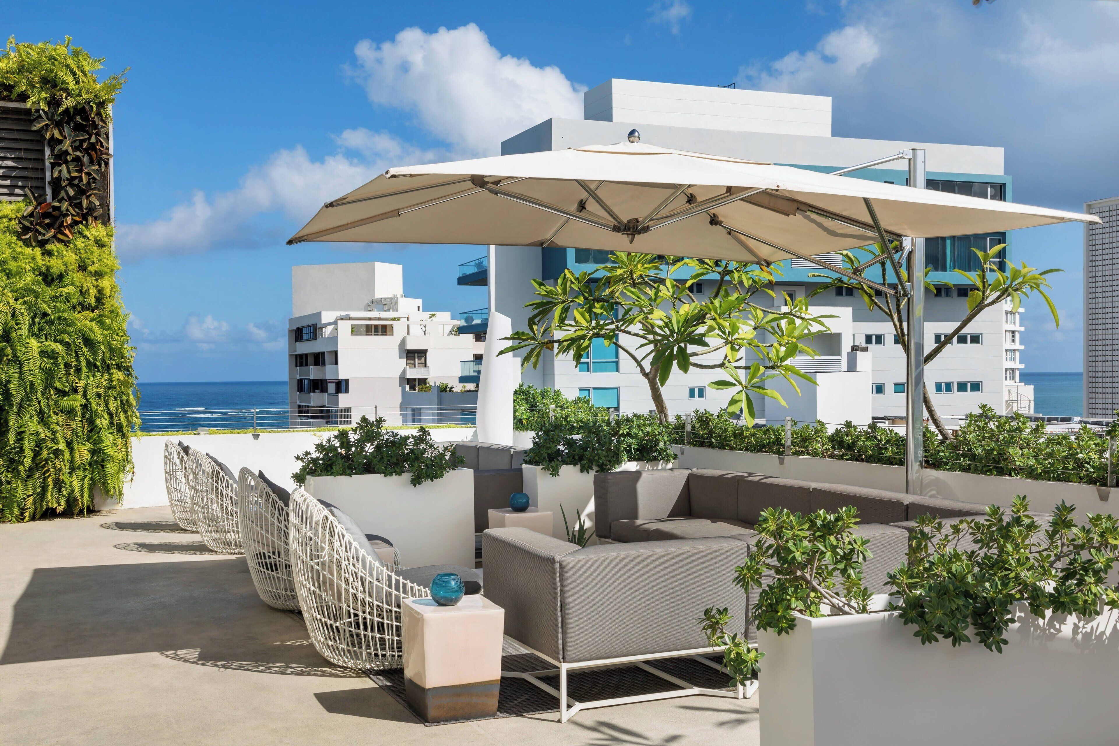 Eine Dachterrasse mit Außenbestuhlung, einem großen Sonnenschirm und Blick auf das Meer und die Gebäude.
