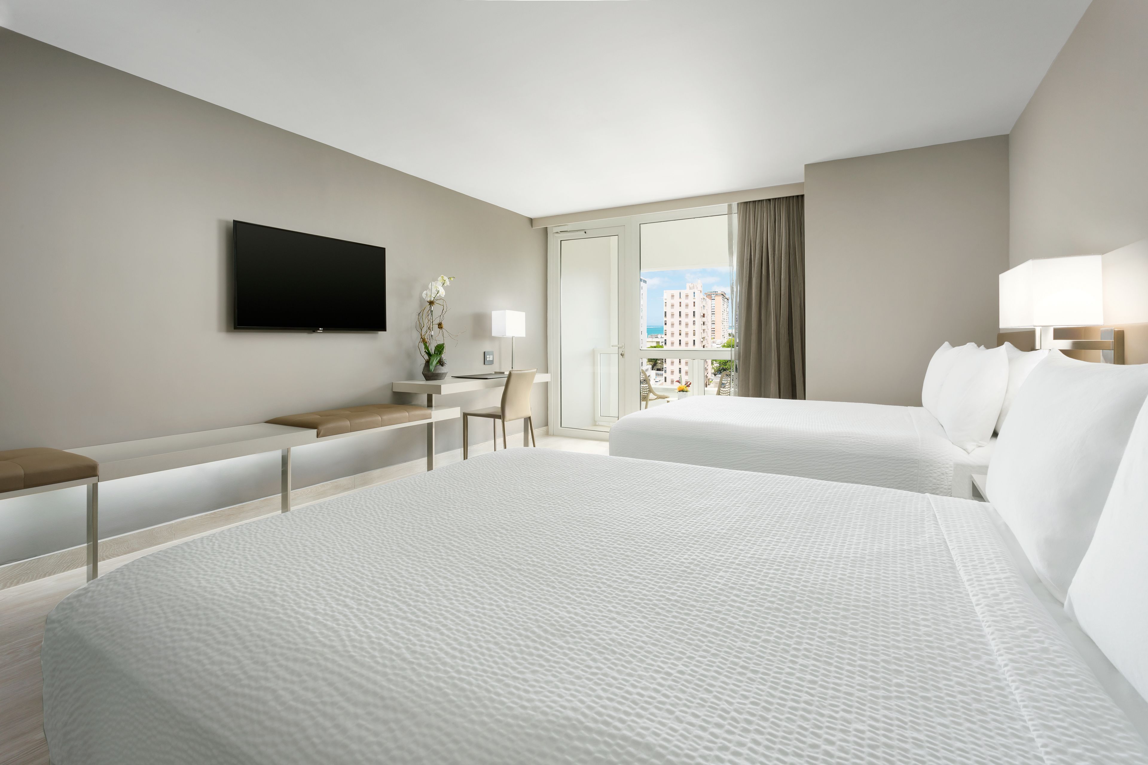 A hotel room with two beds, a desk, a TV, and a large window with a view of buildings.
