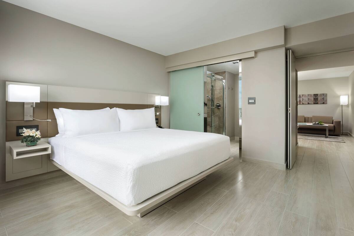 Ein modernes Hotelzimmer mit einem großen Bett, Nachttischen inklusive Lampen, einer Dusche mit Glastrennwand und Blick auf den angrenzenden Wohnbereich.