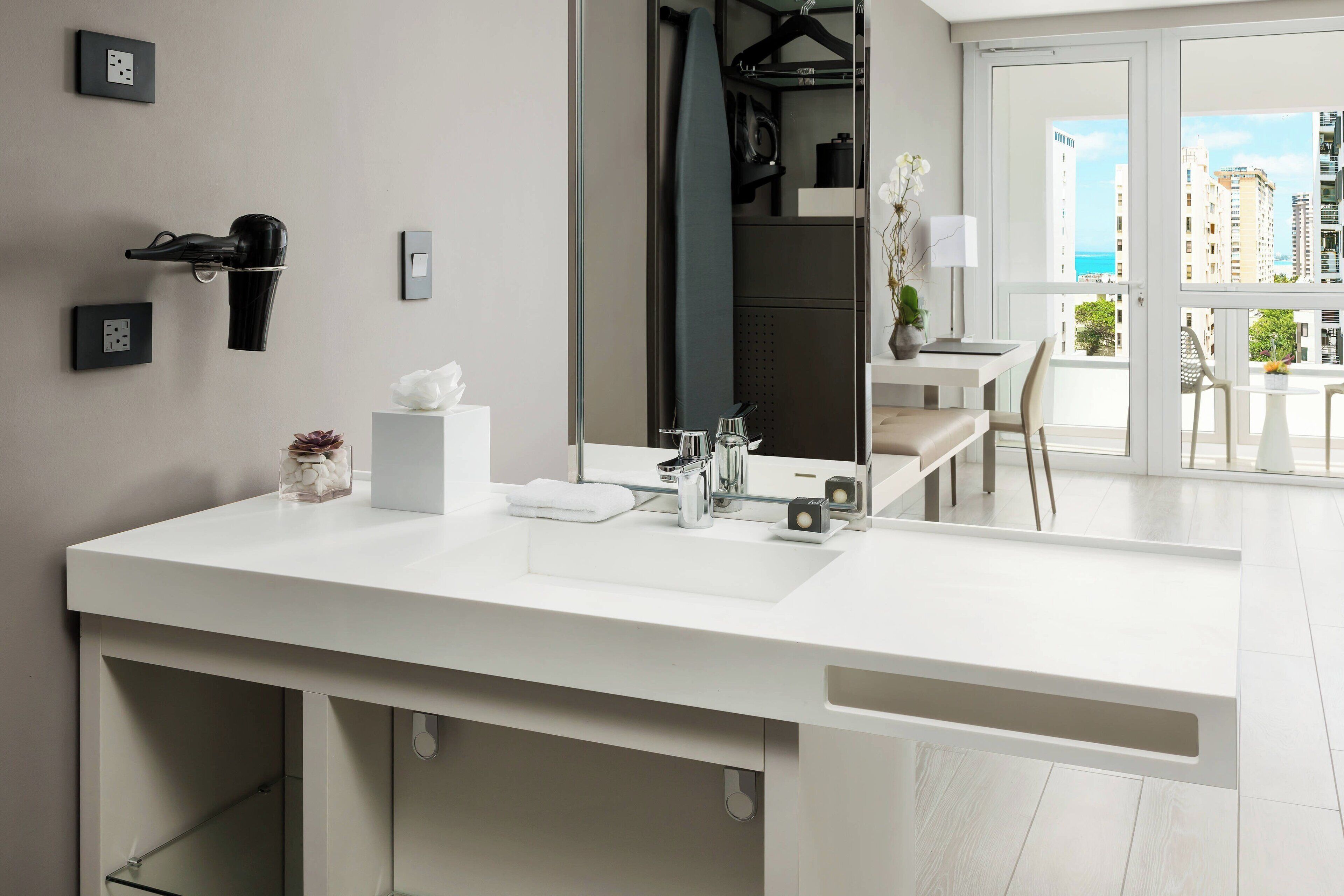 Un baño moderno con un espejo grande, lavamanos blanco y vistas de la ciudad a través de la puerta de cristal.