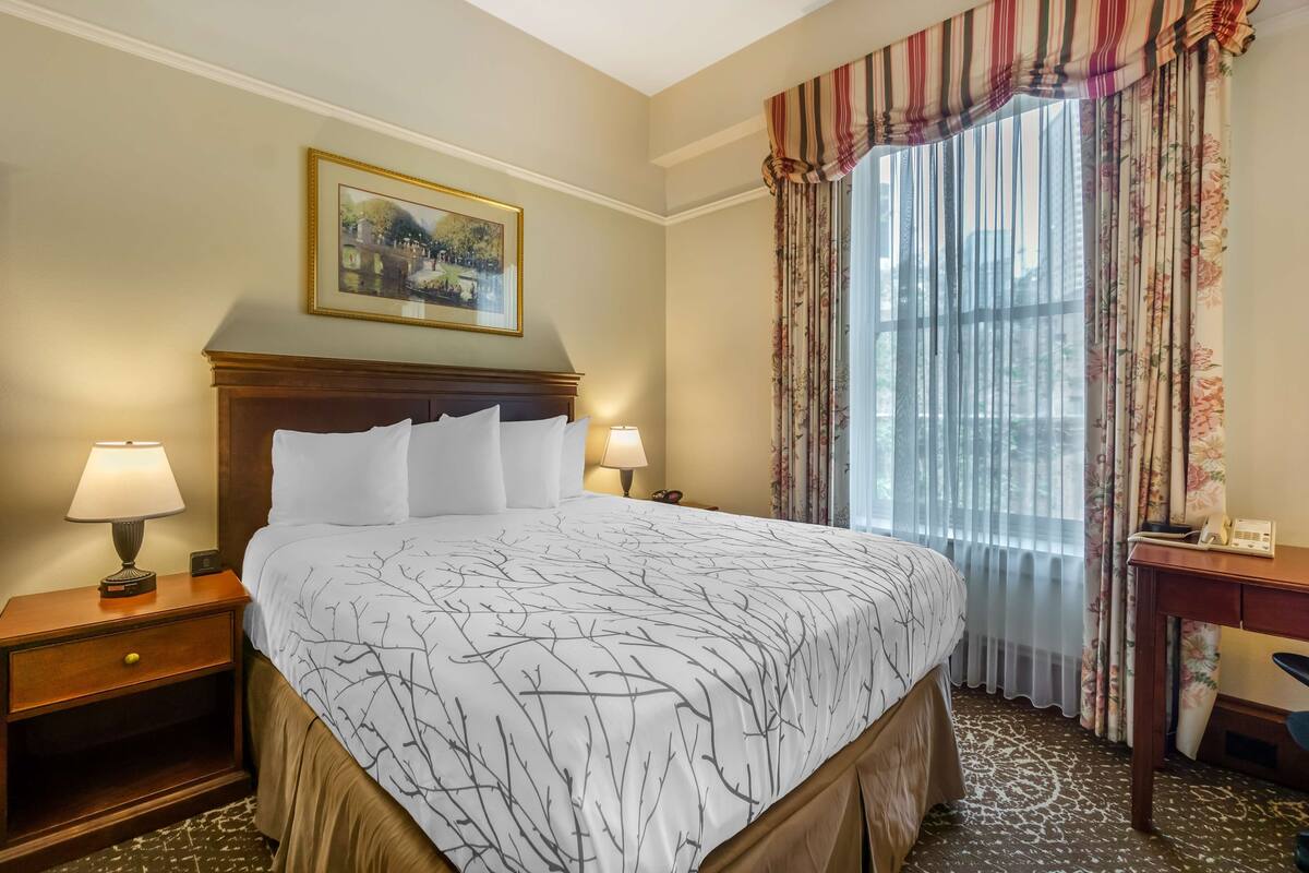 Un cuarto de hotel con una cama grande, dos lámparas de noche, una mesita de noche, un escritorio, una silla, una ventana con cortinas y un cuadro en la pared.