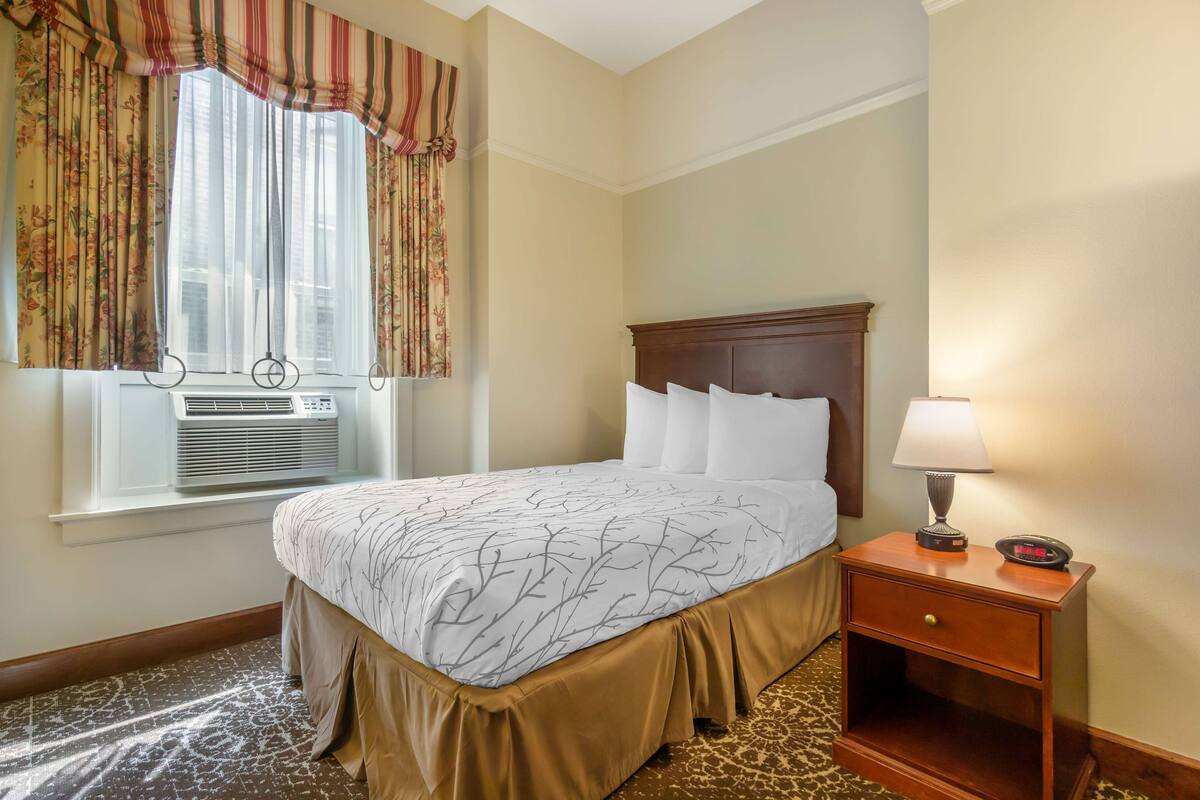 Una habitación de hotel con cama, mesita de noche, lámpara y ventana con cortinas.