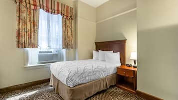 Una habitación de hotel con cama, mesita de noche, lámpara, ventana con cortinas y aire acondicionado.
