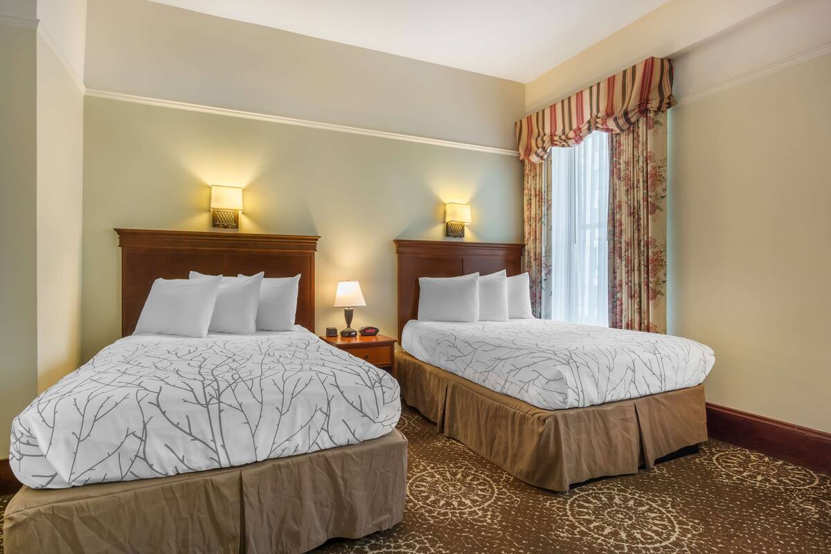 Dos camas dobles con cabeceras y pies de cama a juego, una mesita de noche con lámpara y una ventana con cortinas florales.