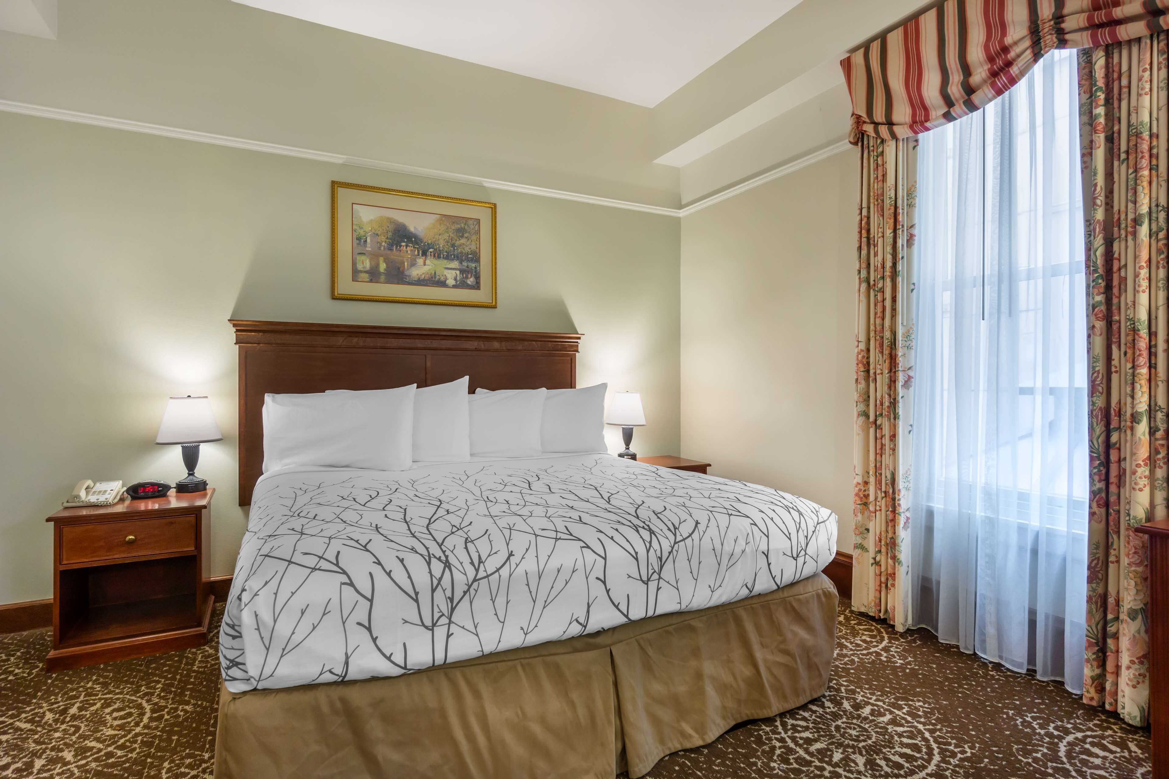 Een hotelkamer met een groot bed, twee nachtkastjes met lampen, een schilderij aan de muur en een raam met bloemen gordijnen.