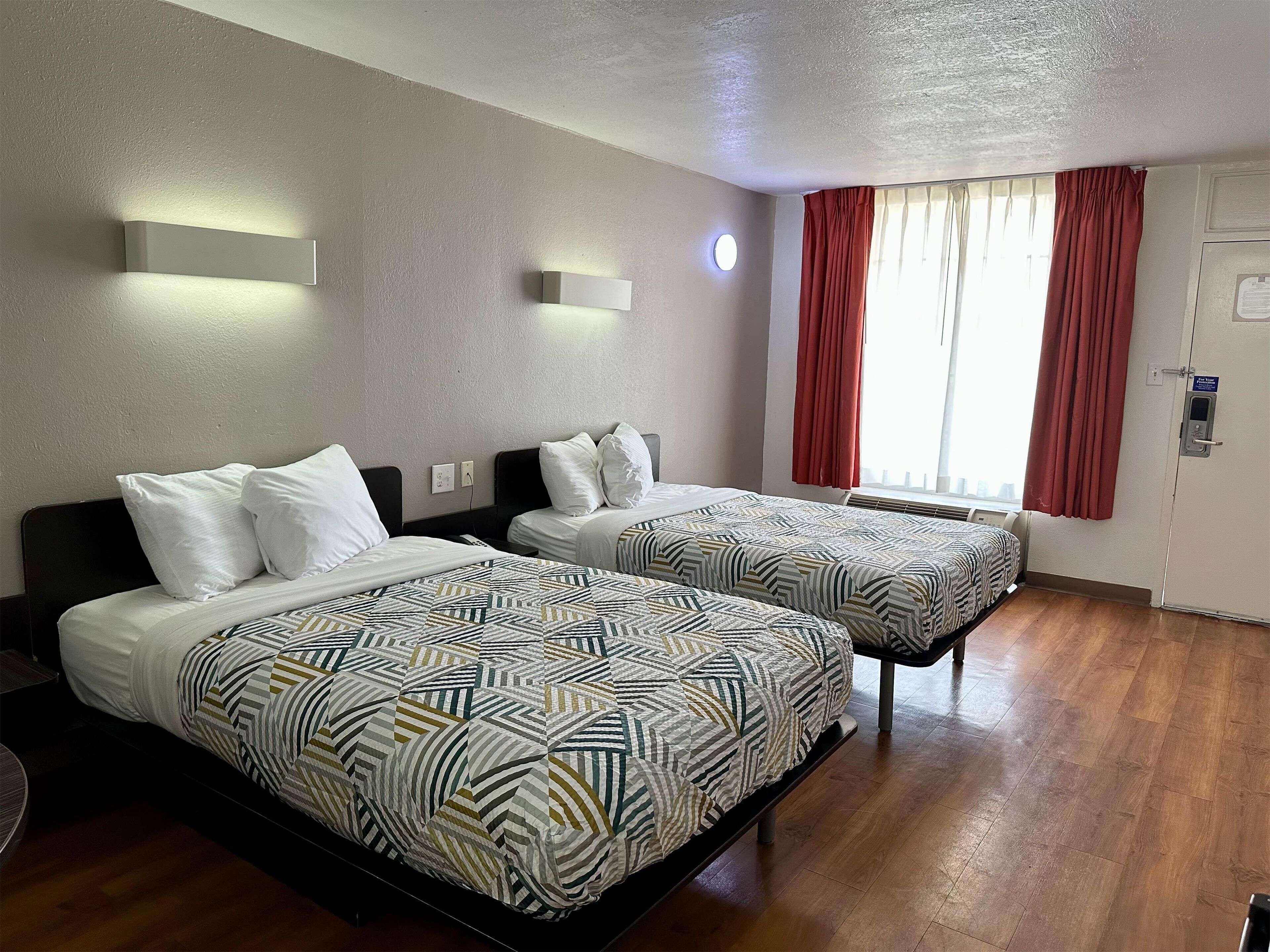 A hotel room with two beds, a window with red curtains, a wall-mounted light, and a door with a sign.