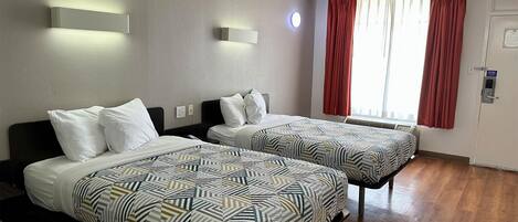A hotel room with two beds, a window with red curtains, a wall-mounted light, and a door with a sign.