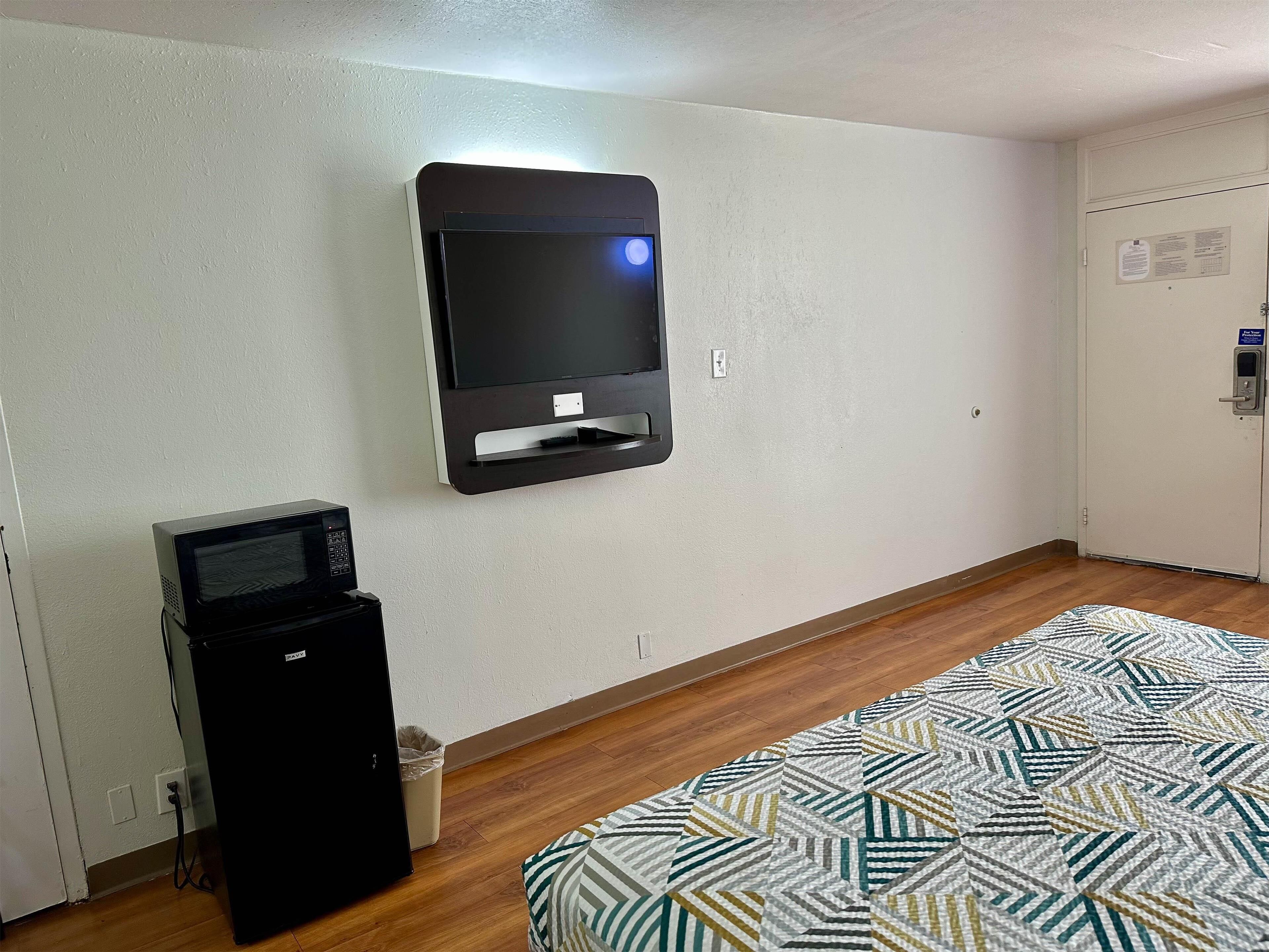 A room with a flat-screen TV mounted on the wall, a small refrigerator, a patterned rug, and a door with a lock and a keycard reader.