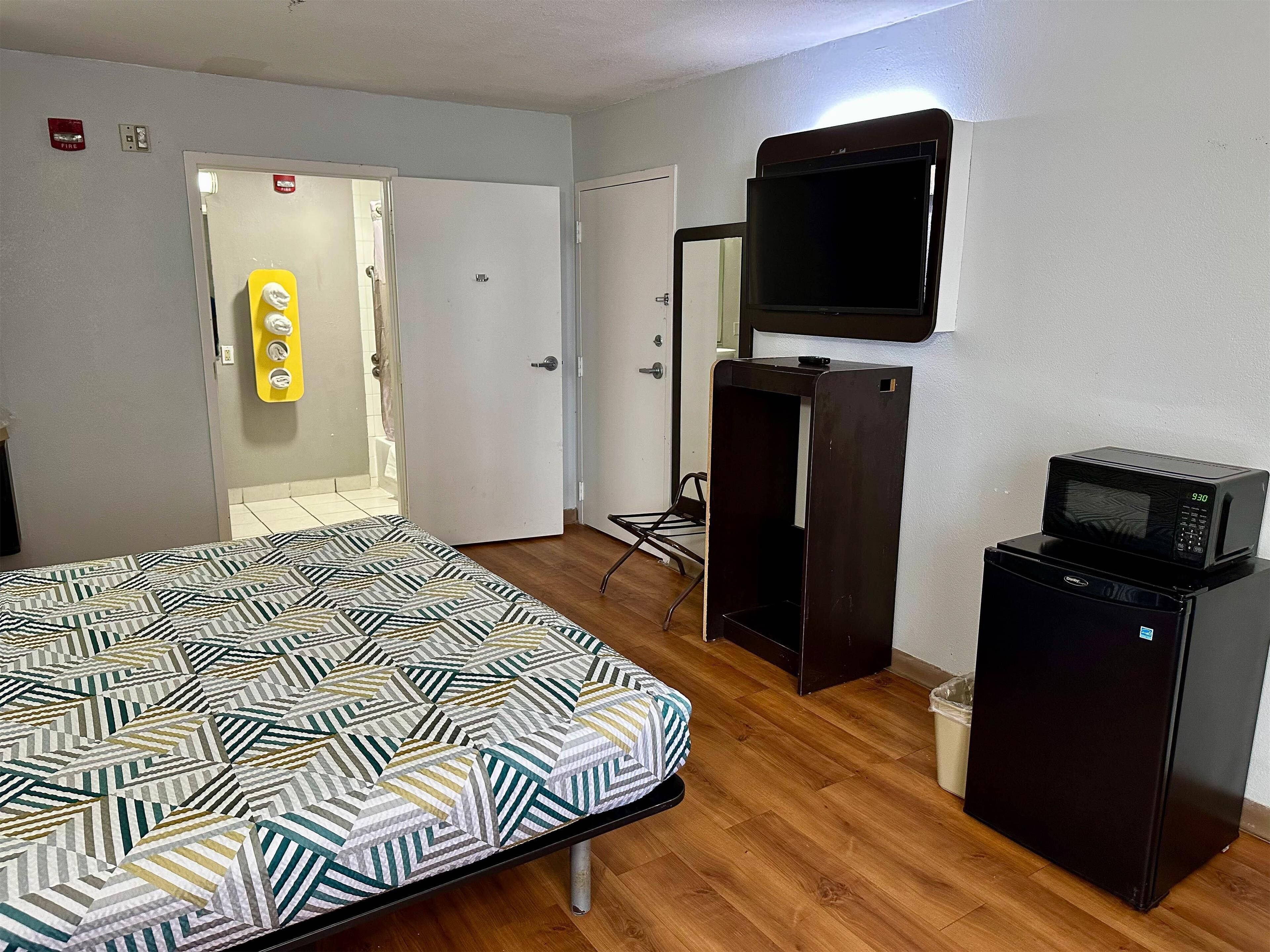 A hotel room with a bed, a TV mounted on the wall, a small refrigerator, and a door leading to a bathroom.