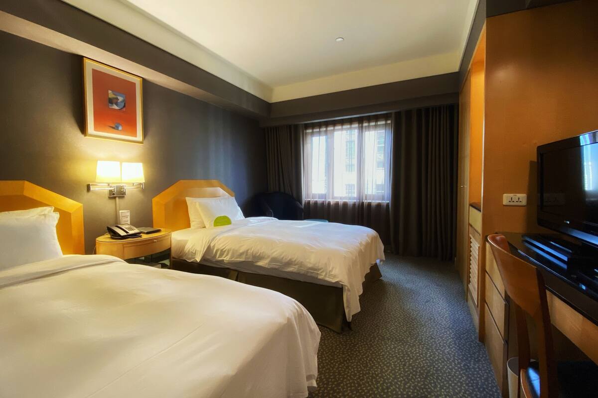 A hotel room with two beds, a television, a desk with a telephone, and a window with curtains.