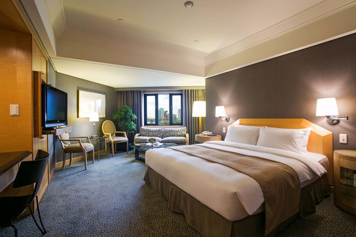 A hotel room with a large bed, a desk with a computer, a chair, a sofa, and a window with a city view.
