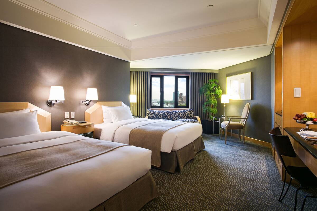 A hotel room with two beds, a desk, a chair, a window with a view, and a plant.