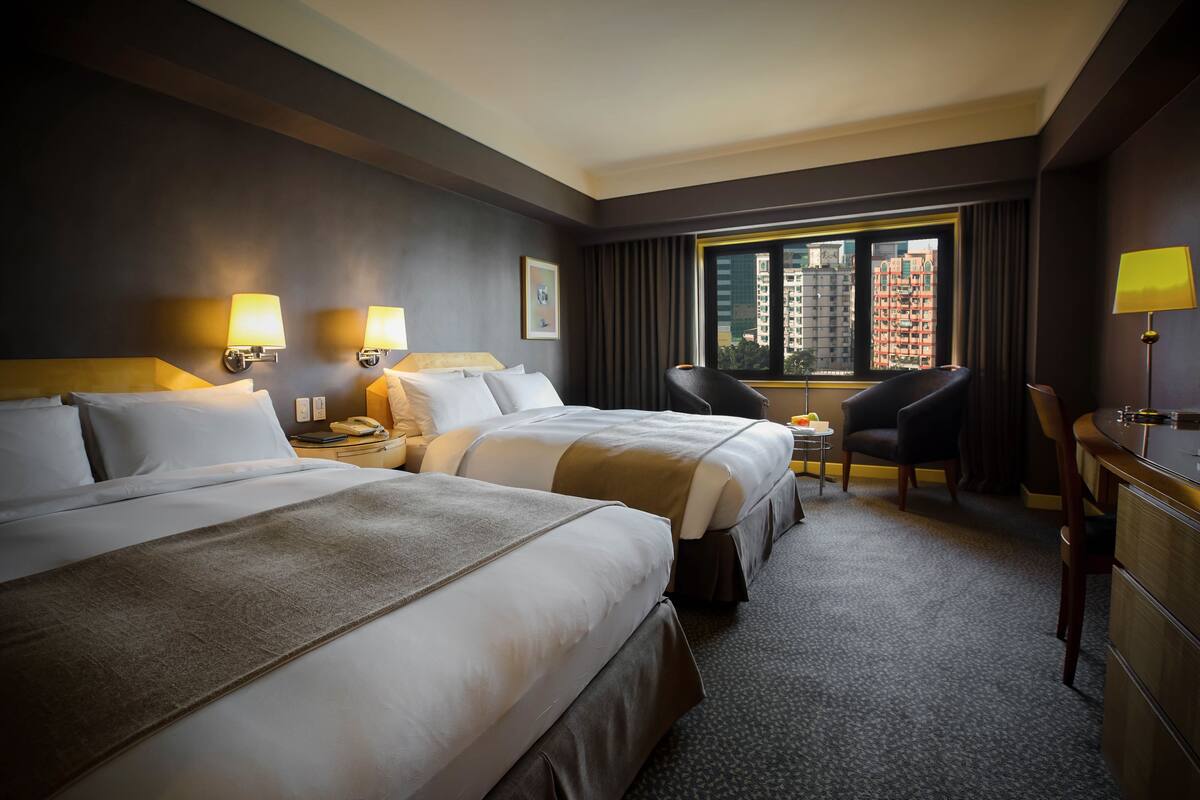 A hotel room with two beds, a desk, a chair, and a view of the cityscape.
