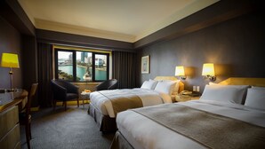 Une chambre d’hôtel avec deux lits, un bureau, une chaise et une vue sur le paysage urbain, visible à travers une grande fenêtre.