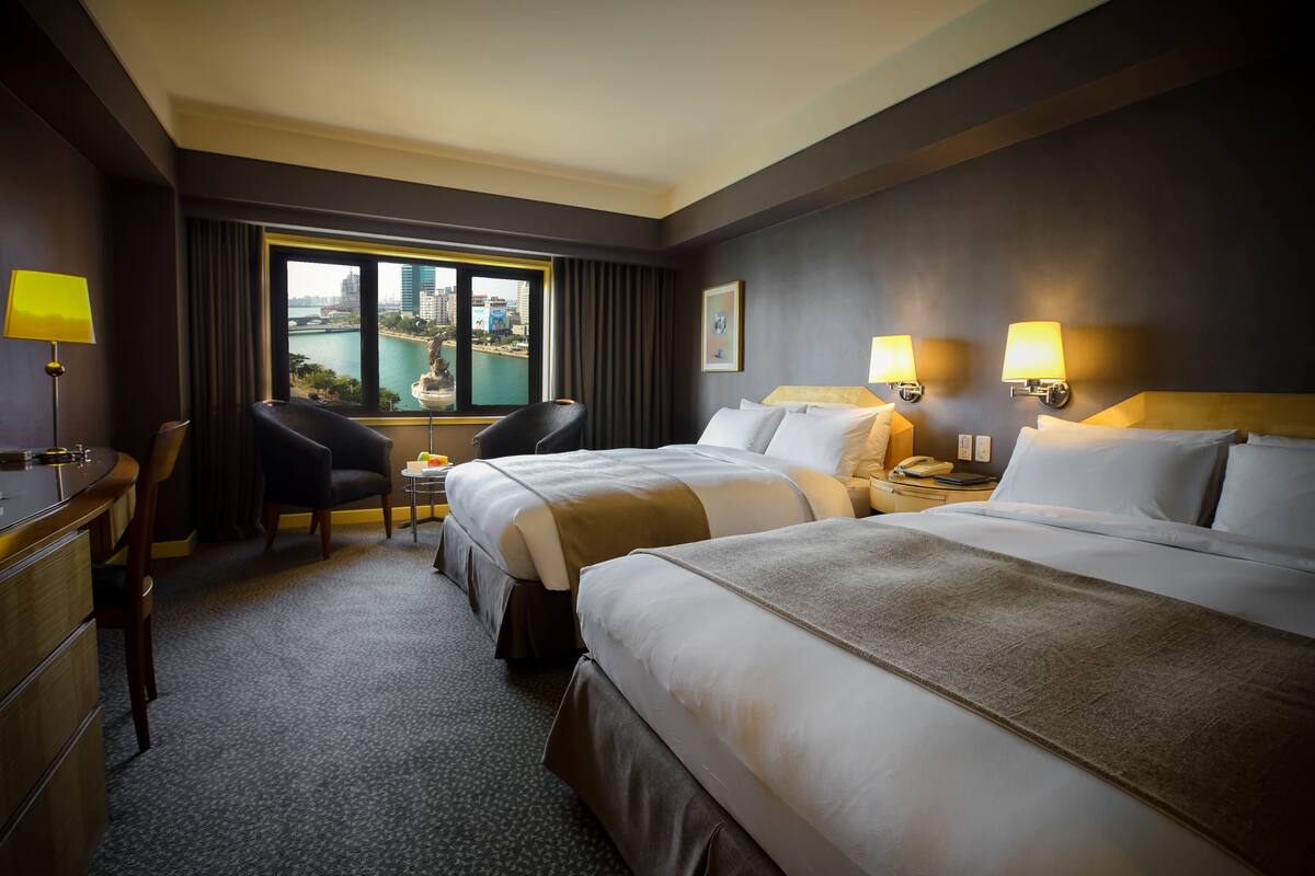A hotel room with two beds, a desk, a chair, and a view of a cityscape through a large window.
