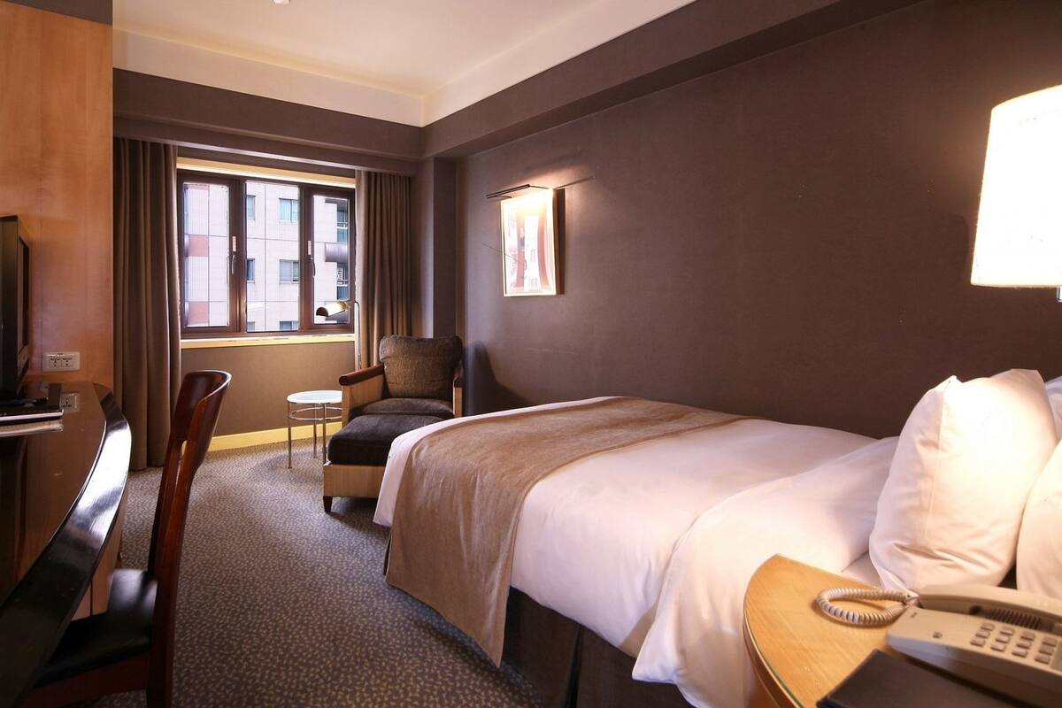 A hotel room with a bed, a desk, a chair, a small table, and a window with curtains.