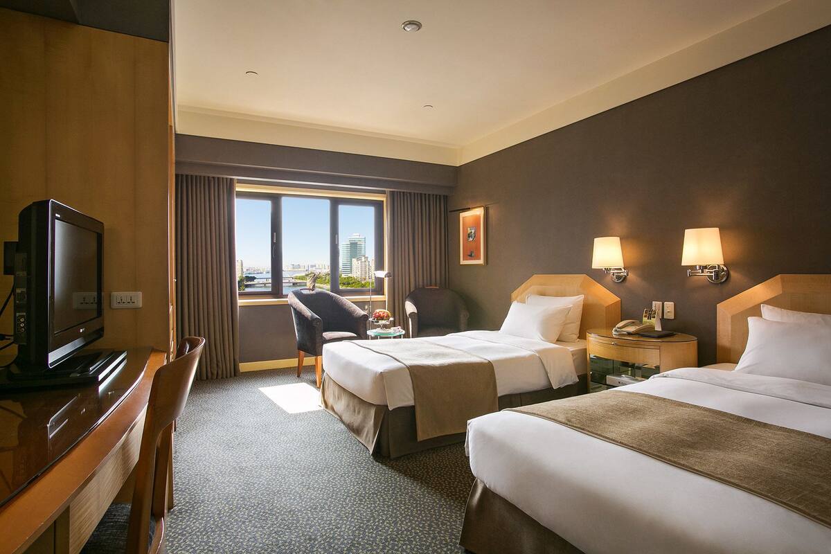 A hotel room with two beds, a desk, a television, and a view of the city.