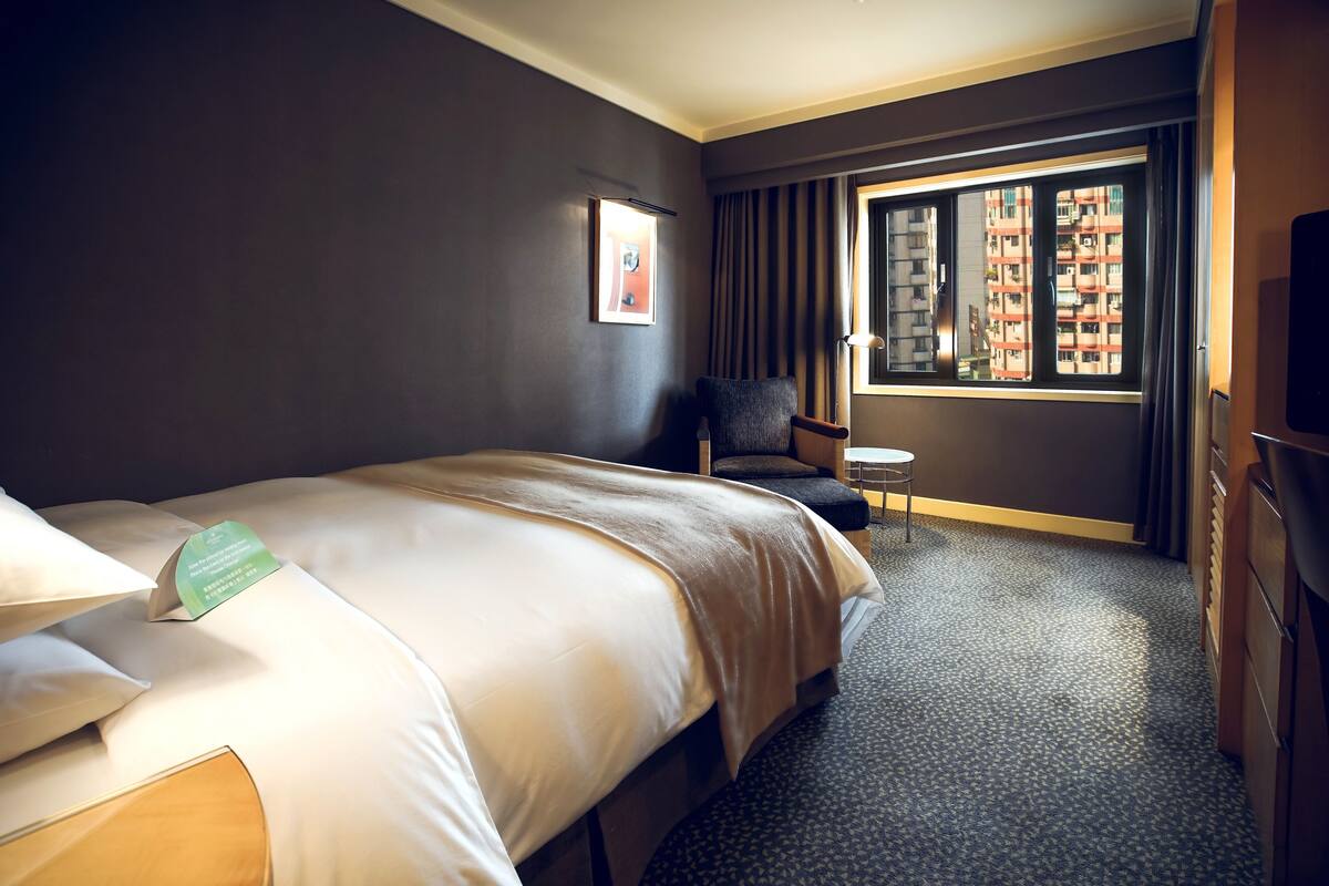 A hotel room with a large bed, a chair, a small table, and a window with a view of buildings.