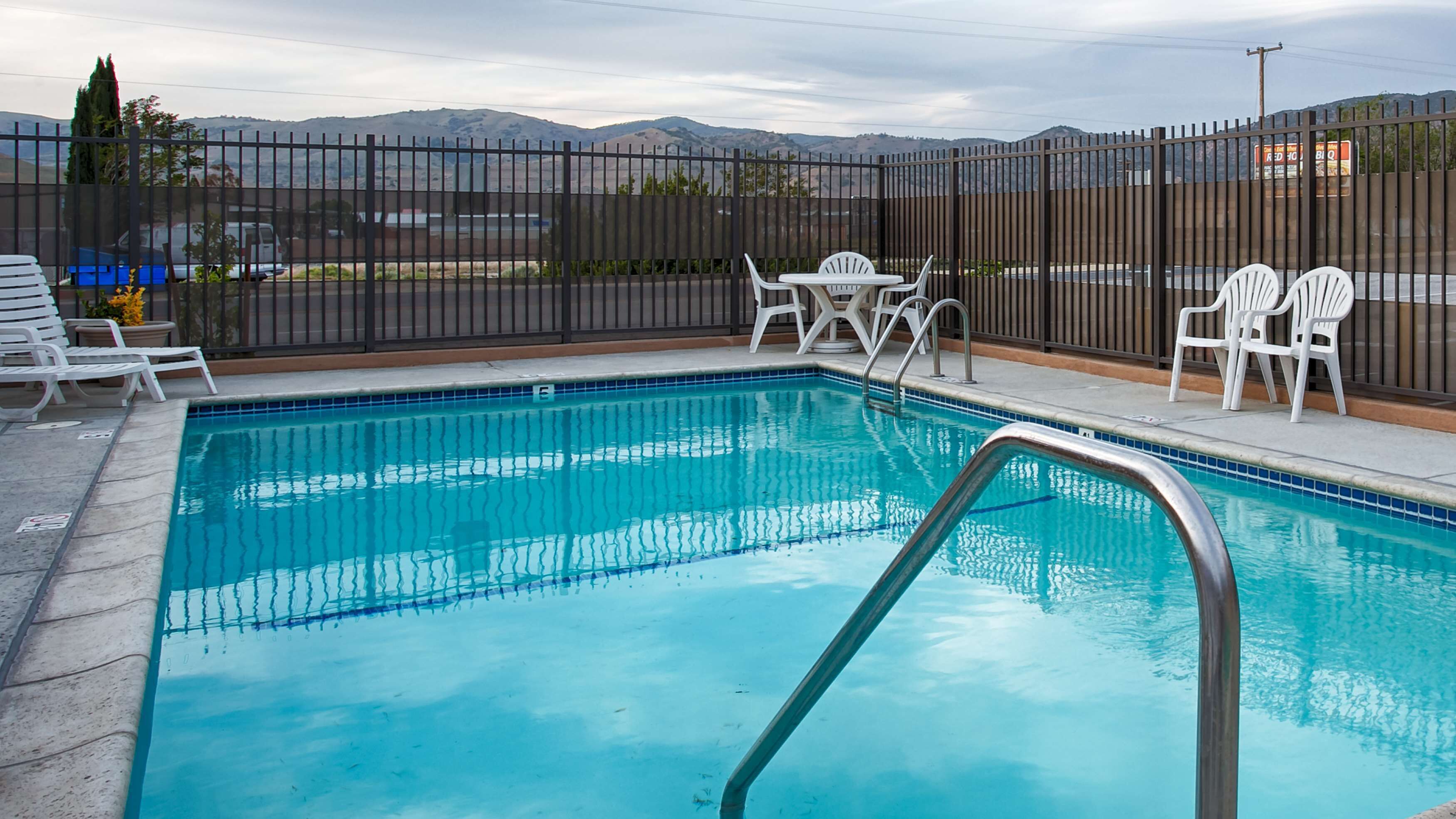 A rectangular pool with clear blue water, surrounded by a black fence. There are white plastic chairs and a table near the pool.