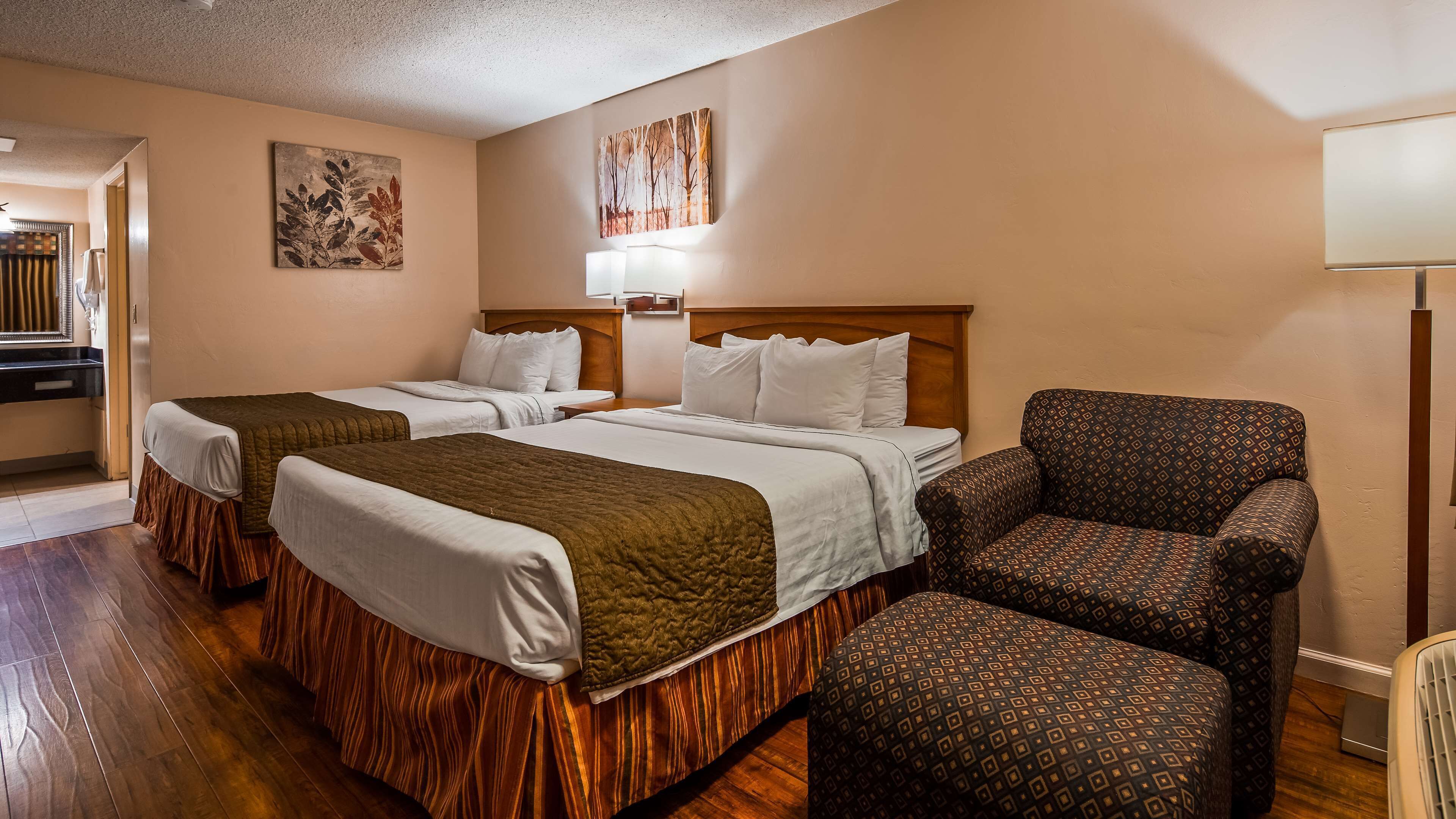 A hotel room with two beds, a patterned armchair, a nightstand, and artwork on the walls.