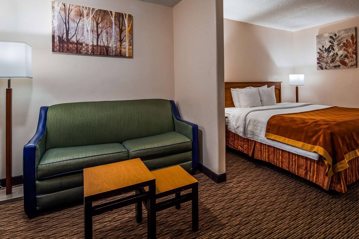 A hotel room with a green sofa, a wooden table, a bed with a brown bedspread, and two wall-mounted lamps.