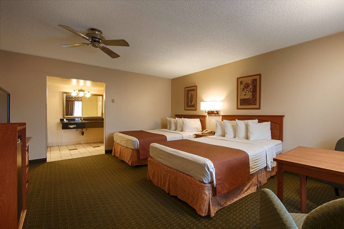A hotel room with two beds, a desk, a chair, a ceiling fan, a bathroom with a mirror, and two wall-mounted lamps.