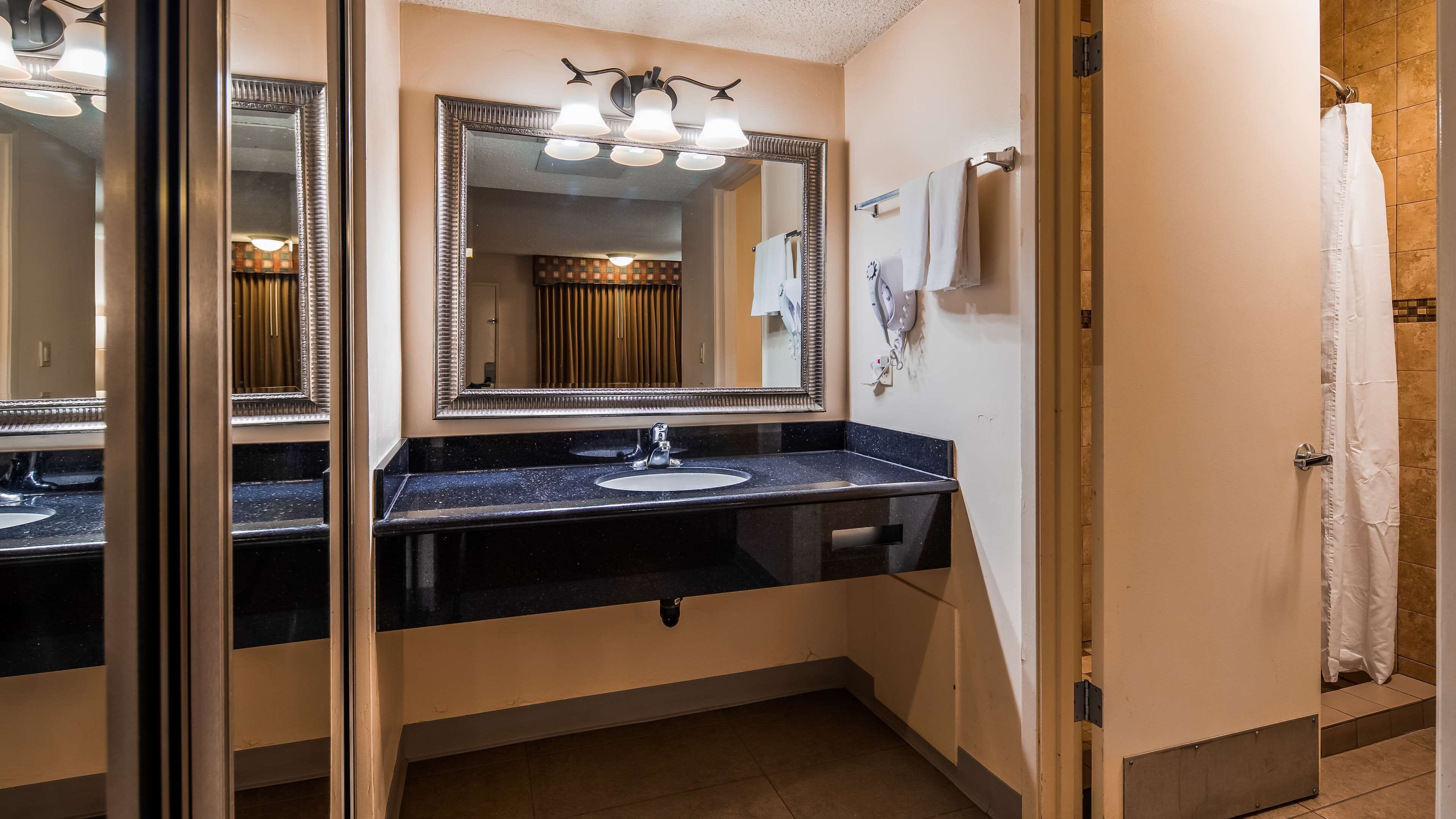 A bathroom with a black countertop, a white sink, and a large mirror with three lights. There is a towel rack and a shower curtain visible.