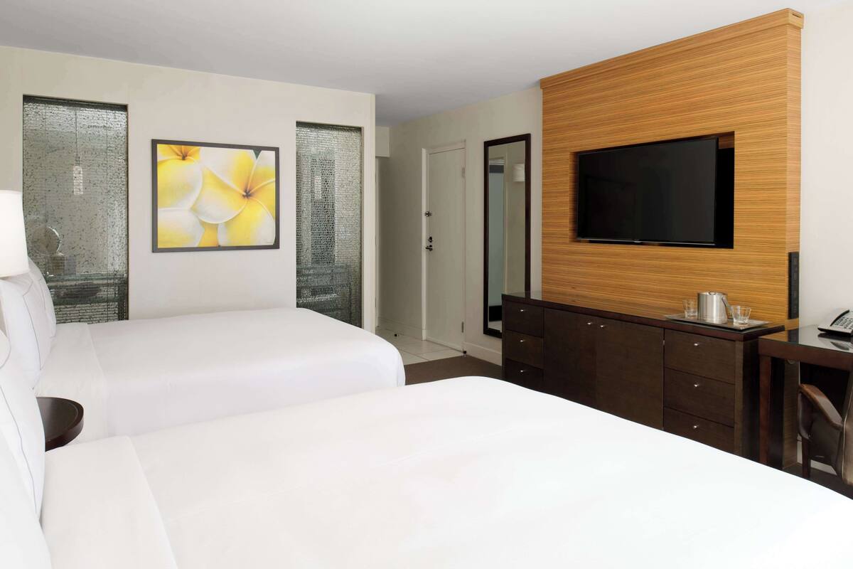 A hotel room with two beds, a TV mounted on a wooden panel, a desk with a telephone, and a door leading to another room.