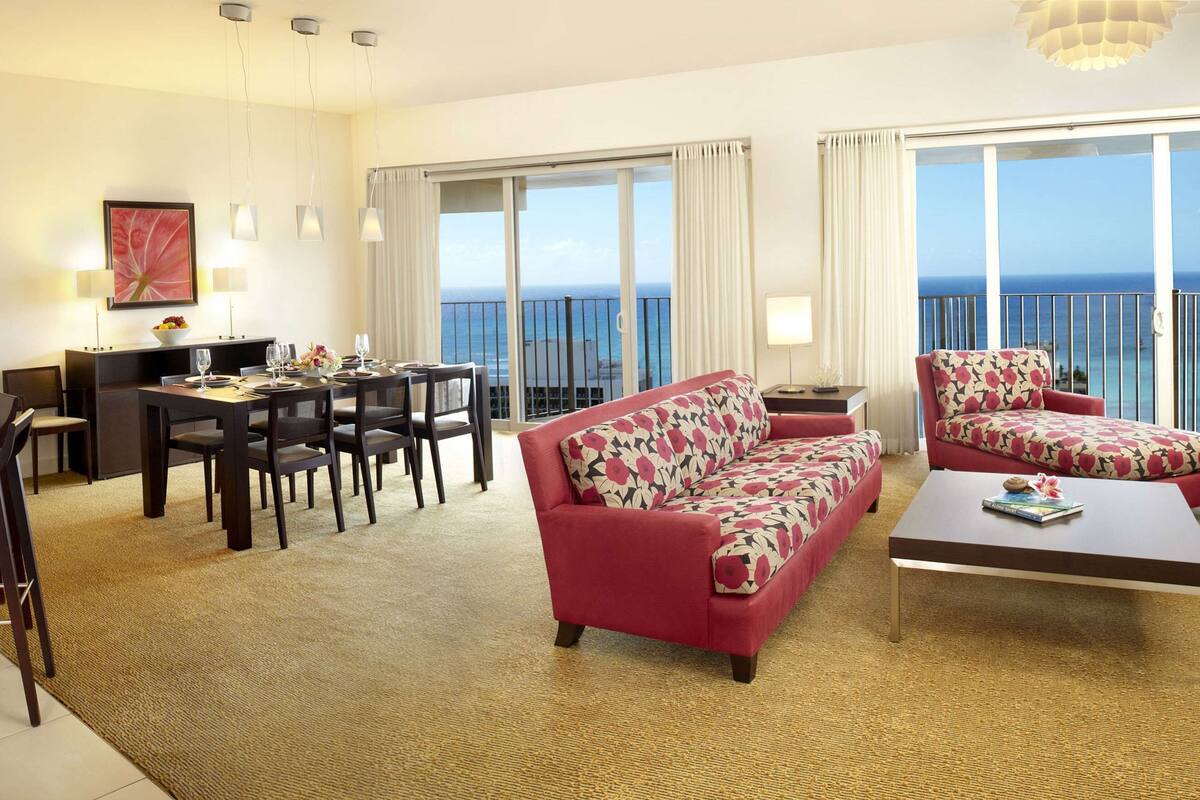 A modern hotel room with a dining area, a sofa, and a balcony with a view of the ocean.