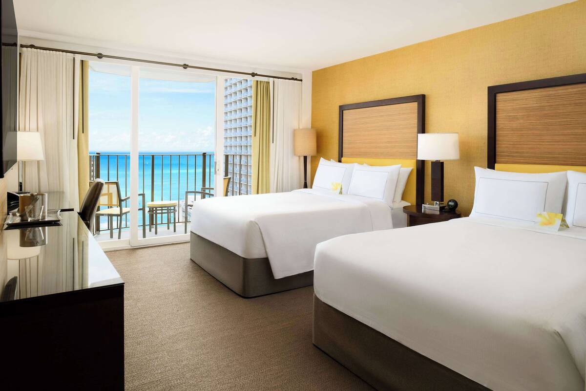 A hotel room with two beds, a desk, a chair, and a balcony with a view of the ocean.