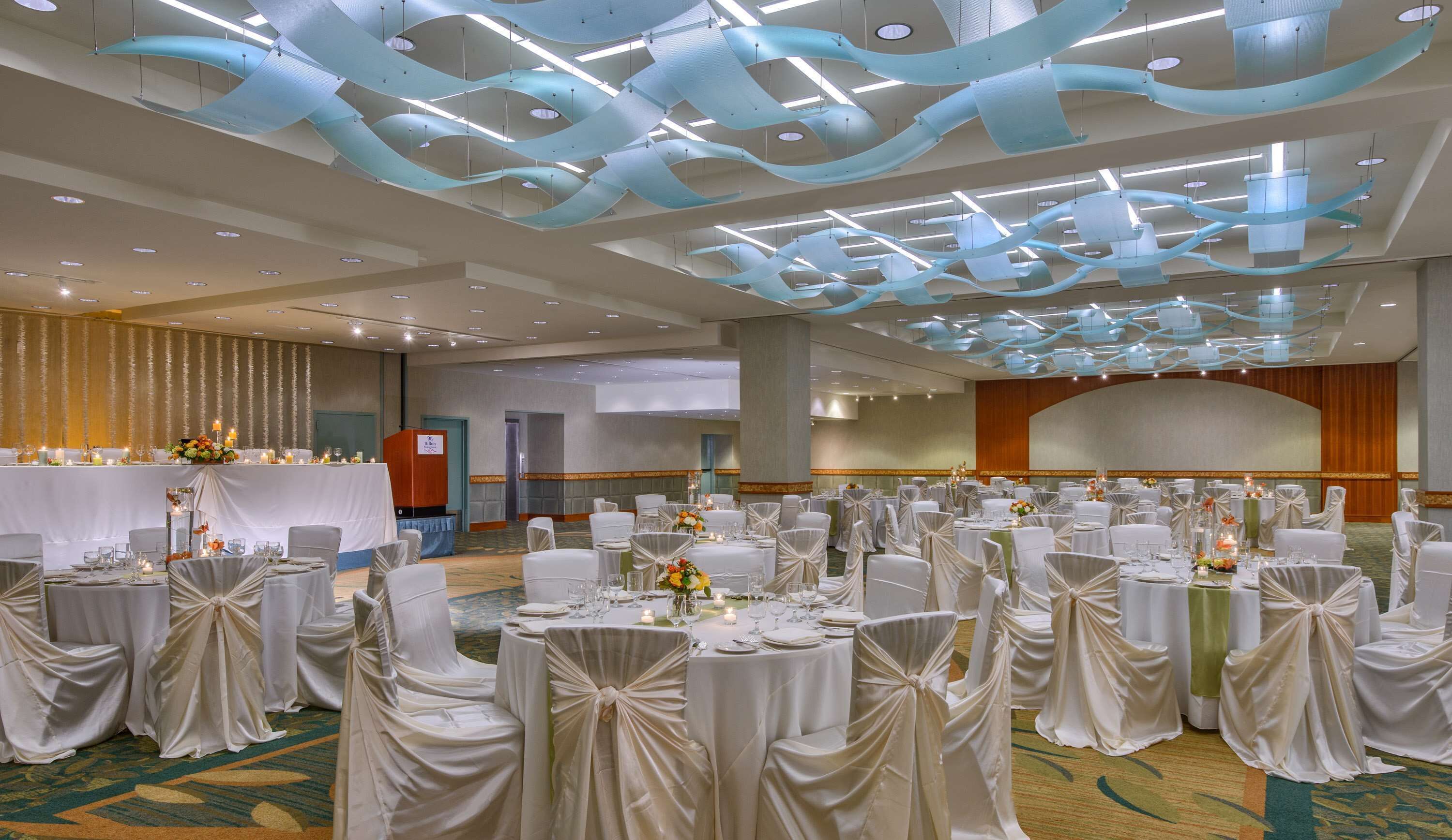 Un salón de banquetes con mesas redondas, manteles blancos y sillas con cintas blancas.