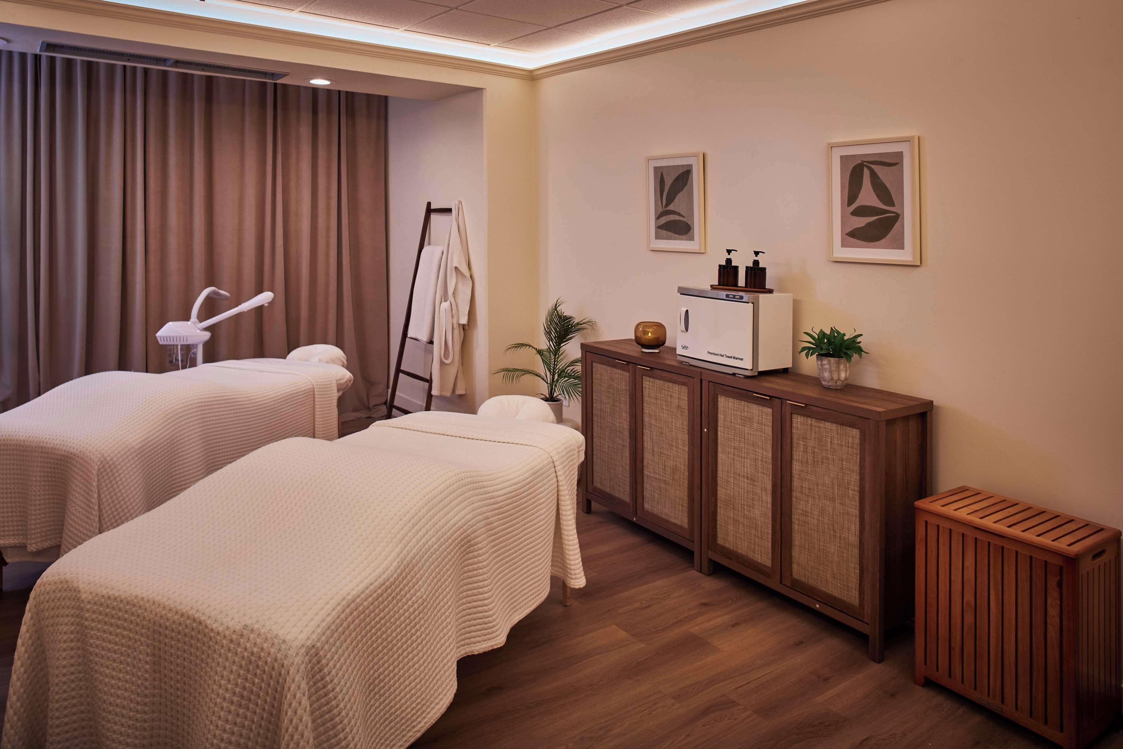 A spa room with two massage tables, a cabinet with toiletries, and a potted plant.