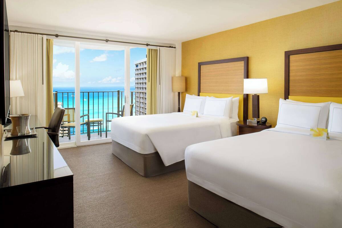 A hotel room with two beds, a balcony with a view of the ocean, and a desk with a lamp.