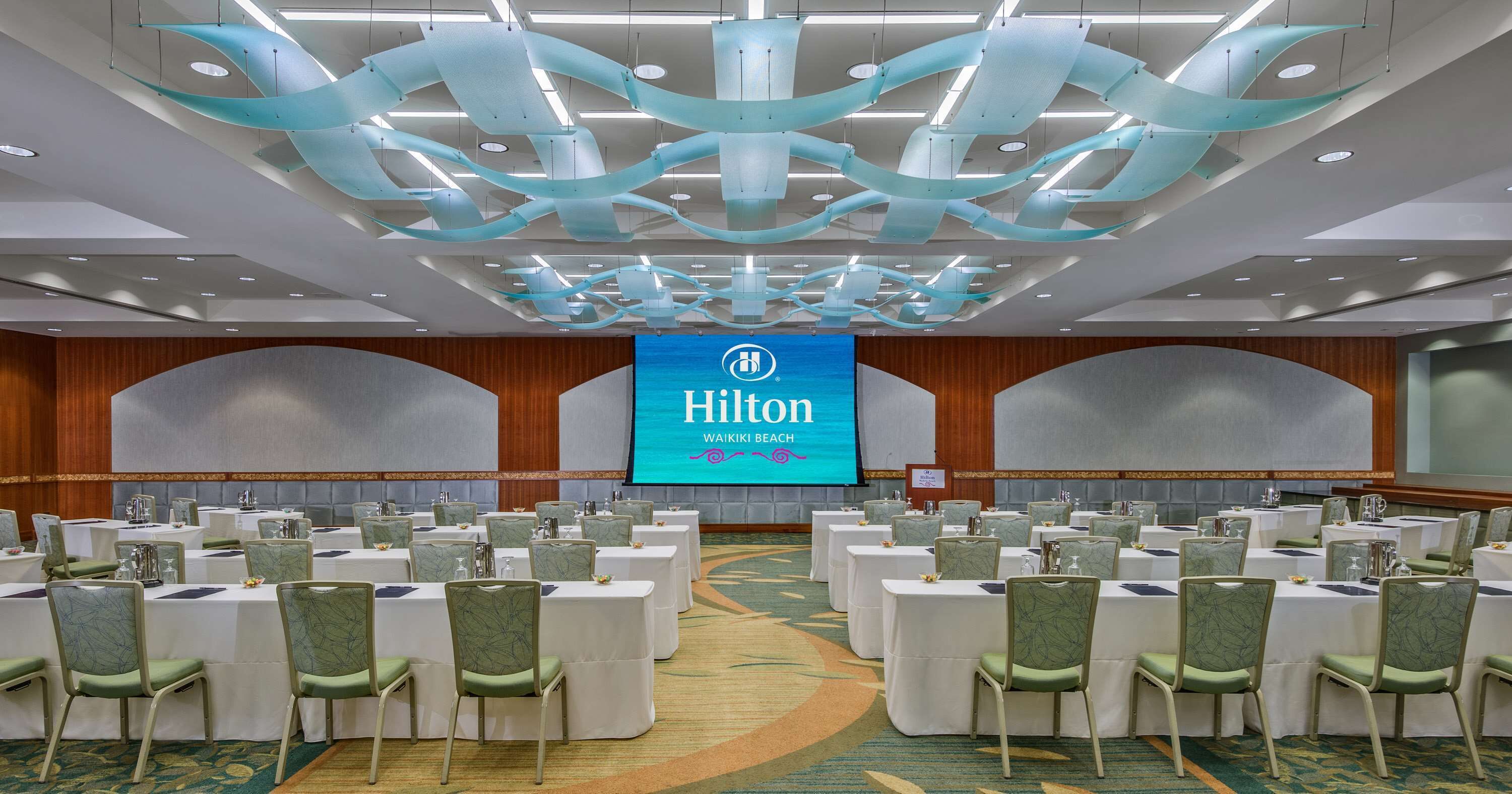 Ein Konferenzraum im Hilton Waikiki Beach mit mehreren Tischen und Stühlen, einem großen Bildschirm mit dem Hilton-Logo und einem modernen Deckenstil.