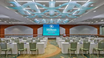Ein Konferenzraum im Hilton Waikiki Beach mit mehreren Tischen und Stühlen, einem großen Bildschirm mit dem Hilton-Logo und einem modernen Deckenstil.