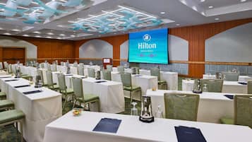 A conference room with rows of tables and chairs, a large screen displaying 'Hilton Waikiki Beach', and a podium with a microphone.
