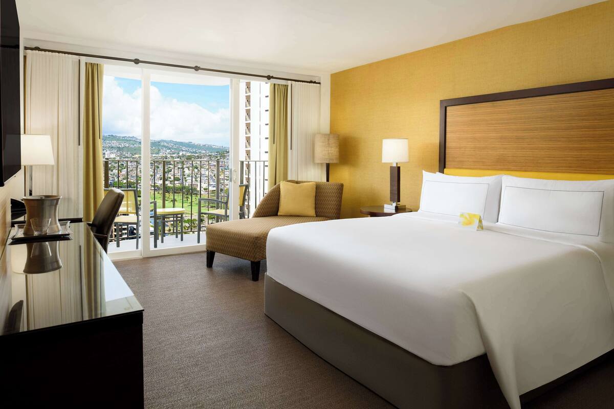 A hotel room with a large bed, a desk with a chair, a balcony with a view of the city, and a window with curtains.