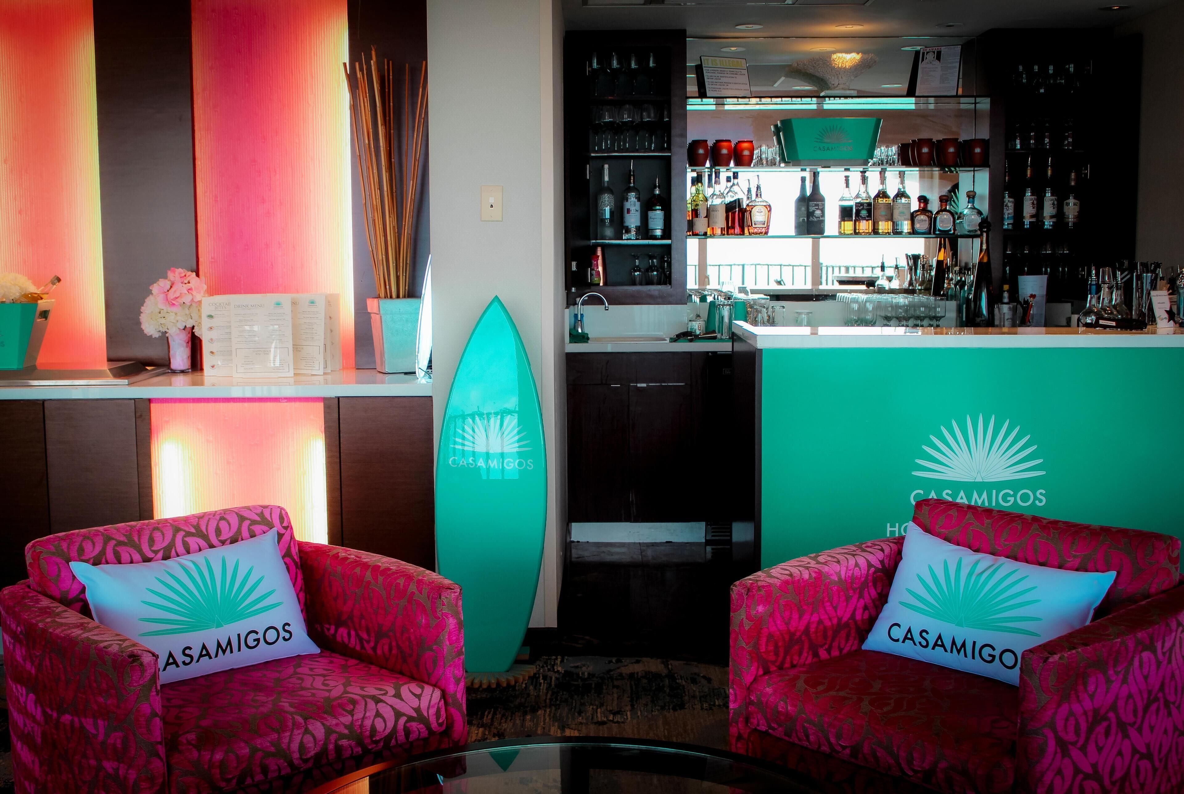 Een bar met een toonbank, flessenrekken en een surfboard. Twee stoelen met 'CASAMIGOS' kussens staan op de voorgrond.