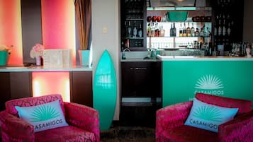 Een bar met een toonbank, flessenrekken en een surfboard. Twee stoelen met 'CASAMIGOS' kussens staan op de voorgrond.