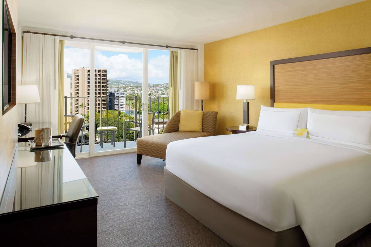 A hotel room with a large bed, a desk with a chair, two lamps, and a view of a cityscape through large windows.