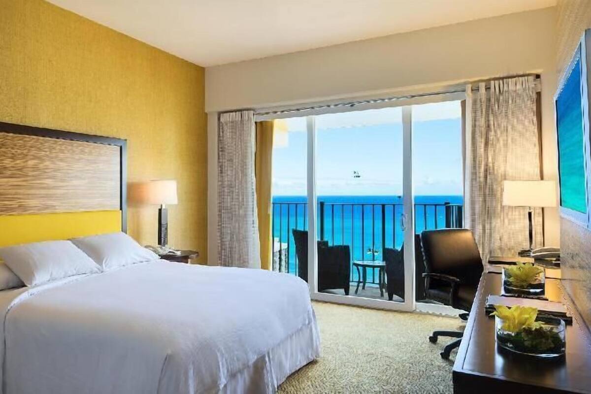 A hotel room with a bed, a desk, a chair, and a balcony with a view of the sea.