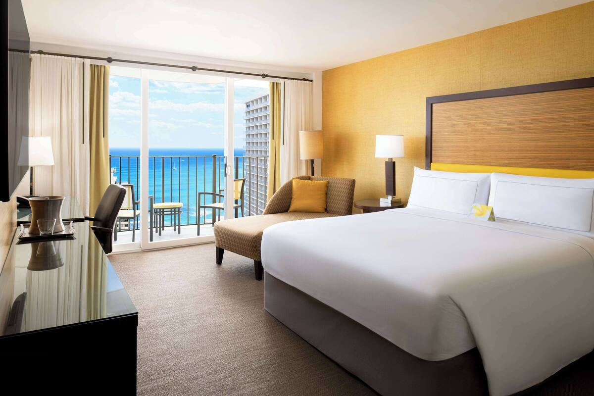A hotel room with a large bed, a desk, a chair, and a balcony with a view of the ocean.